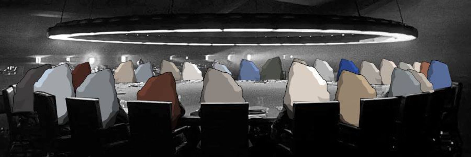 A council of colored boulders seated in chairs around a boardroom table beneath a ring light